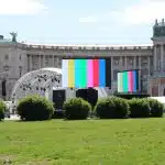 Digital screens displaying colorful test bars are set up on a green lawn in front of a historic building with arched windows, stone statues, and trimmed bushes under a clear blue sky. The modern screens contrast with the classical architecture, creating a striking visual scene.