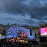 Outdoor concert at dusk in front of historic building with crowd watching illuminated stage and large screen displaying colorful graphics against dramatic cloudy sky.