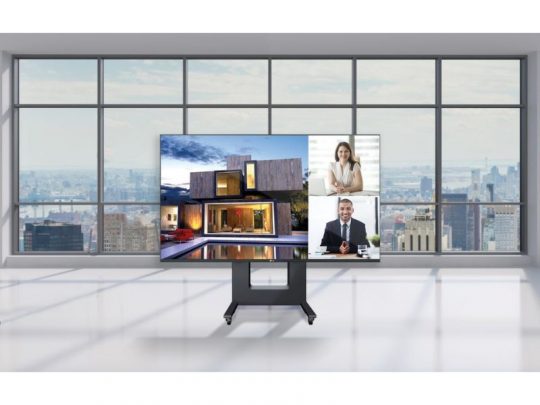 Samsung Neo QLED TV on a slim stand in a bright modern living room with floor-to-ceiling windows; the screen shows a video call with two people and an image of a contemporary house, with city skyline visible outside.