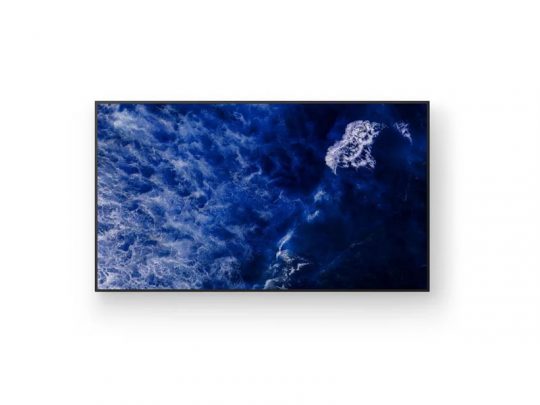 Samsung 4K UHD flat-screen TV mounted on a plain white wall, displaying a vivid aerial image of deep blue ocean waves and white sea foam.