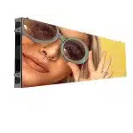 Rectangular LED screen displaying a close-up image of a person with light hair, round sunglasses, a nose ring, and multiple rings on their fingers, holding their hands up in front of a bright yellow background. Ideal for digital signage or modern advertising displays.