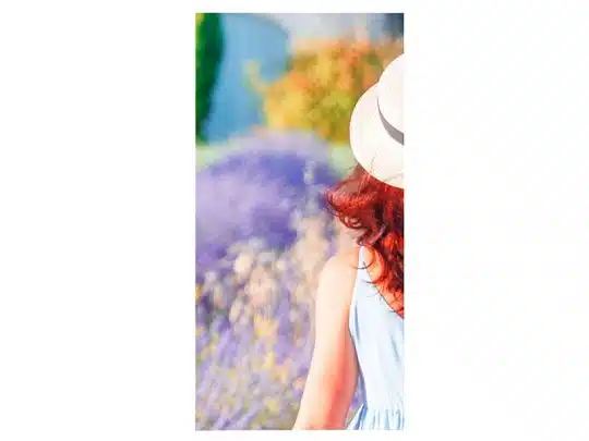 A woman with red hair, wearing a light blue summer dress and a straw hat, stands outdoors with her back to the camera amid vibrant blurred flowers and green foliage in bright sunlight—ideal for showcasing spring or summer fashion.