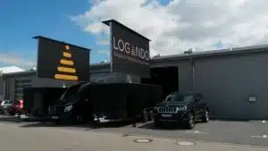Zwei schwarze Fahrzeuge stehen vor einem grauen Industriegebäude mit großen "LOGONDO Display & Media Solutions GmbH"-Schildern. Vor dem Gebäude ist der LED Trailer 15m² - 5,12m x 2,88m V:LED VSF6 LED Screen gemietet und sichtbar aufgebaut. Der Himmel ist teilweise bewölkt.