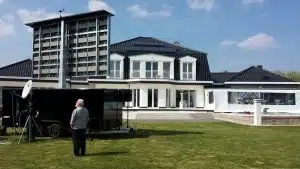 Modernes Einfamilienhaus mit dunklen Dachziegeln und Solarpanels; davor steht ein hoher Stapel elektronischer Geräte. Auf der Wiese neben einem schwarzen Anhänger mit Satellitenschüssel steht ein Mann. Perfekt zum Kaufen oder Mieten - großzügiges, energieeffizientes Haus bei sonnigem Wetter und blauem Himmel.