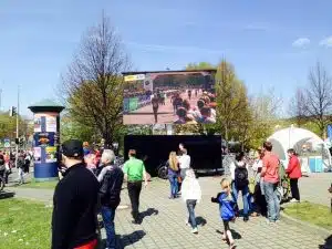 Großbildleinwand im Park für Outdoor-Events - Menschen unterschiedlichen Alters versammeln sich an einem sonnigen Tag vor einer großen LED-Leinwand, um eine Veranstaltung zu verfolgen. Im Hintergrund sind Bäume und weiße Zelte zu sehen. Die mobile Großleinwand ist für den Kauf oder die Miete geeignet, ideal für Public Viewing oder Open-Air-Events.