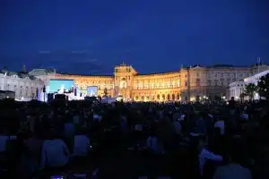 Große Menschenmenge sitzt nachts auf einer Wiese vor einem hell beleuchteten, historischen Gebäude mit hohen Rundbogenfenstern; davor Bühne mit leuchtenden Bildschirmen für öffentliches Open-Air-Event oder Konzert. Die Event-Location ist für Veranstaltungen miet- oder kaufbar.