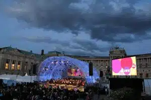 Beleuchtetes Open-Air-Bühnenzelt mit Orchester und großem Publikumsbereich vor einem historischen Gebäude bei Sonnenuntergang; das professionelle Bühnenzelt mit LED-Display ist für Konzerte und Events zu kaufen oder zu mieten, Bühne im Fokus vor malerischer Abendkulisse.