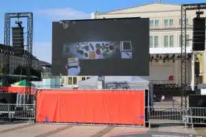 LED Trailer Cabrio 15m² (5,12m x 2,88m) V:LED VSF6 LED Screen zur Vermietung vor einem klassischen Gebäude; der große mobile LED-Bildschirm zeigt eine Top-down-Ansicht von Speisen und einem Notizblock, umgeben von Absperrungen und Event-Ausrüstung.