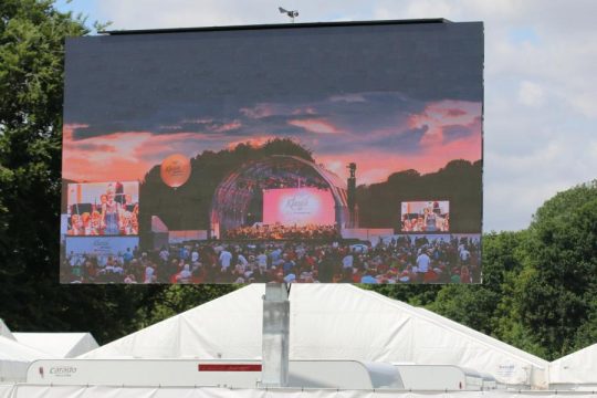 LED Trailer Cabrio 23m² - 6,40m x 3,60m V:LED VSF6 bei Sonnenuntergang: Der mobile LED-Anhänger zeigt ein Live-Konzert über weißen Zelten, umgeben von Bäumen und Wolken im Hintergrund. Ideal zum Kaufen oder Mieten für Events und Großveranstaltungen.