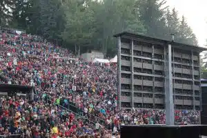Große Menschenmenge sitzt im Freien in einem Amphitheater, umgeben von Bäumen. Im Vordergrund ist ein hoch aufragendes Lautsprechersystem (verfügbar zum Mieten oder Kaufen) und eine Bühne zu sehen. Das Publikum wartet auf den Beginn einer Veranstaltung oder Show.