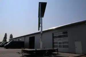 Großes, vertikales LED-Elektronik-Display (Verkehrszeichen) auf Anhänger vor grauer Industriehalle mit Solaranlage und schwarzen Transportern bei Sonnenschein - perfekt zum Mieten oder Kaufen.