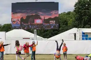 Kinder in orangefarbenen Warnwesten spielen und machen Radschläge auf einer Wiese vor einer großen Outdoor-Leinwand (Miete oder Kauf möglich), die bei Sonnenuntergang ein Konzert zeigt; im Hintergrund sind weiße Zelte und Bäume zu sehen.