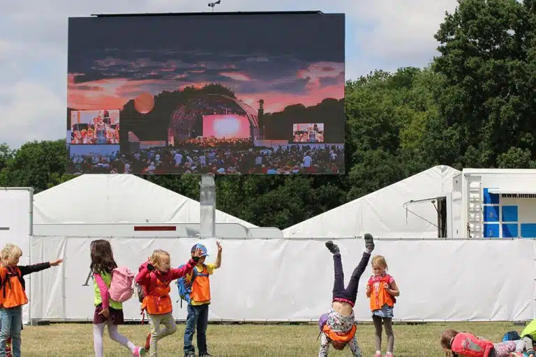 Kinder in orangefarbenen Warnwesten spielen und machen Radschläge auf einer Wiese vor einer großen Outdoor-Leinwand (Miete oder Kauf möglich), die bei Sonnenuntergang ein Konzert zeigt; im Hintergrund sind weiße Zelte und Bäume zu sehen.