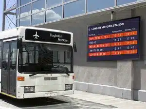 Weißer Shuttlebus steht vor einem Gebäude mit einem 75 Zoll Outdoor LCD Display - Samsung OH75F zur Miete. Das Display zeigt internationale Abflüge nach New York, Dubai und London Heathrow für den London Victoria Station an. Ideal für Outdoor-Werbung und Fahrgastinformationen.