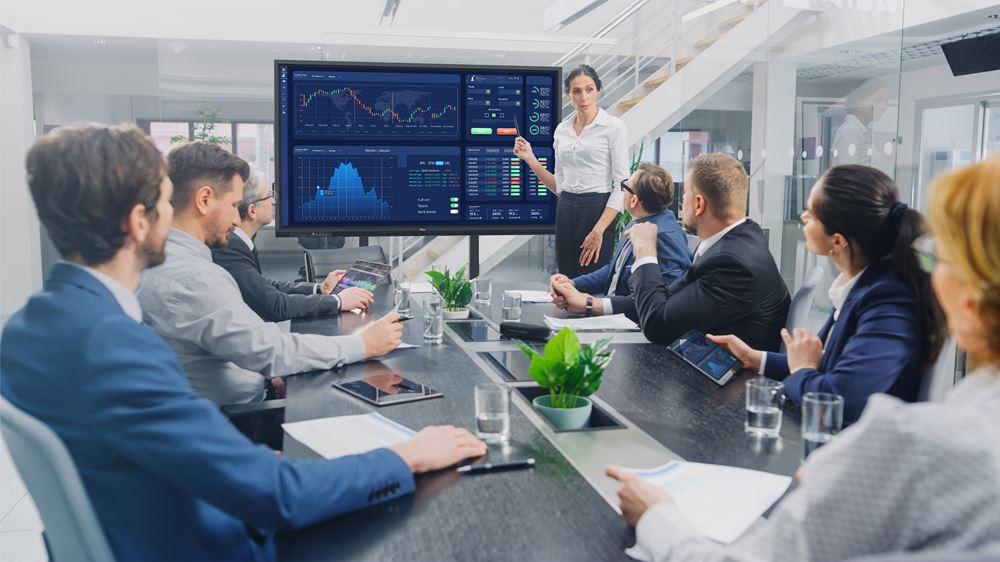 Eine Frau präsentiert Mieten- und Kaufen-Trends auf dem 86 Zoll UHD Multi Touch Display - Optoma 3862RK (Neuware). Das große interaktive Whiteboard zeigt Diagramme und Grafiken, während mehrere Geschäftsleute an einem modernen Konferenztisch im Büro sitzen.