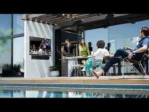 Vier Personen entspannen auf einer sonnigen Terrasse mit bequemen Sitzmöbeln und schauen ein Basketballspiel auf dem 75 Zoll Outdoor 4K QLED-TV - Samsung The Terrace. Der wetterfeste Fernseher steht am Pool vor großen Glas-Schiebetüren - ideal zum Mieten oder Kaufen für Garten und Terrasse.