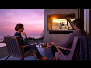 Zwei Personen sitzen bei Sonnenuntergang auf einer Terrasse und schauen einen gemieteten 75 Zoll Outdoor 4K QLED-TV - Samsung The Terrace. Auf dem Bildschirm ist ein Paar am Strand zu sehen, während hinter der Terrasse Stadtlichter leuchten. Auf dem Tisch stehen Getränke; der wetterfeste TV eignet sich ideal für Outdoor-Events.