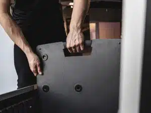 Person im schwarzen Shirt hebt mit beiden Händen ein Gravity TLS 431 B Designstativ für PTZ-Kameras, Licht- und Tontechnik (Mietprodukt) an. Im Hintergrund sind verschwommene Lichter und Möbel eines Innenraums sichtbar.