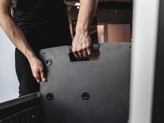 Person im schwarzen Shirt hebt mit beiden Händen ein Gravity TLS 431 B Designstativ für PTZ-Kameras, Licht- und Tontechnik (Mietprodukt) an. Im Hintergrund sind verschwommene Lichter und Möbel eines Innenraums sichtbar.