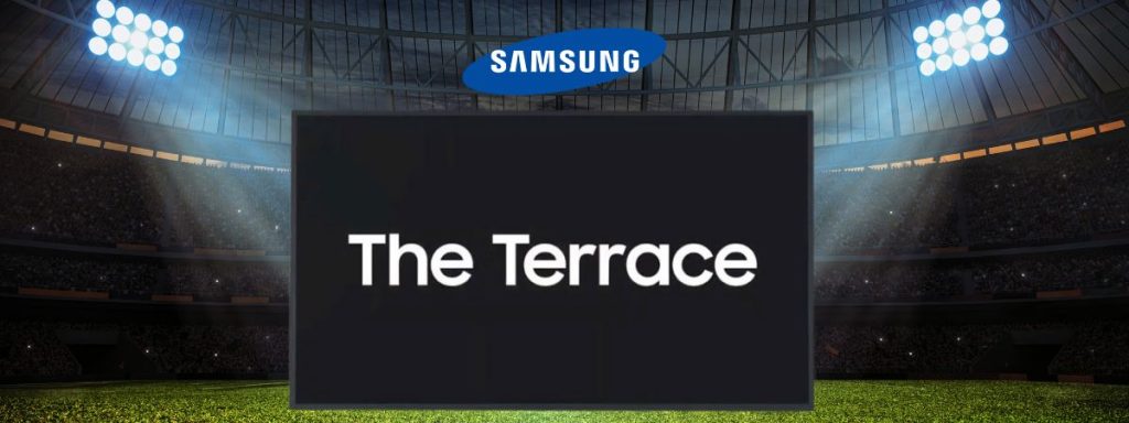 Großer Samsung The Terrace Outdoor-Fernseher mit leuchtendem Bildschirm, auf dem "The Terrace" steht, und sichtbarem Samsung-Logo oben; das Display ist in einem hell beleuchteten Stadion mit Menschenmenge im Hintergrund aufgebaut - ideal für Veranstaltungen, zum Kaufen oder Mieten.