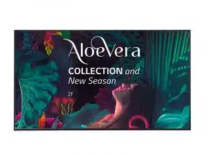 Eine Frau steht umgeben von großen, tropischen Blättern und bunten Pflanzen. Über ihr ist der Schriftzug "Aloe Vera Collection and New Season 2F" zu sehen. Im Hintergrund befindet sich dichtes, farbenfrohes Laub. Das Bild bewirbt die neue Aloe Vera Collection in einer lebendigen Dschungel-Kulisse.