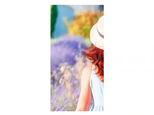 Frau mit roten Haaren trägt einen Sonnenhut und ein hellblaues Kleid; sie ist von hinten zu sehen, spaziert an blühenden Lavendelpflanzen entlang und genießt das sonnige Wetter. Das Bild zeigt eine lebendige, sommerliche Atmosphäre mit unscharfem, farbenfrohem Hintergrund. Ideal als Inspirationsbild für Sommermode oder Lavendel-Feld-Besuche.