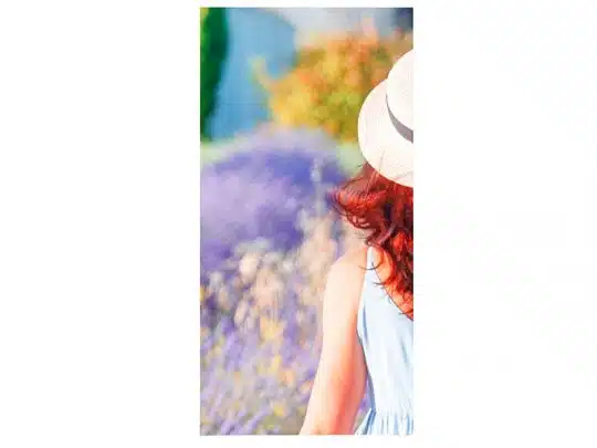 Frau mit roten Haaren trägt einen Strohhut und ein blaues Kleid, steht mit dem Rücken zur Kamera vor einem Feld aus lila und gelben Blumen; sommerliche, farbenfrohe Outdoor-Szene. Ideal als Inspiration für Sommermode mit Strohhut und blauem Damenkleid.