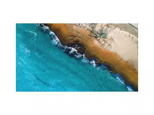 Luftaufnahme von türkisfarbenen Ozeanwellen, die an eine felsige, orangefarbene Küstenlinie prallen; im rechten oberen Bildbereich sind helle Sanddünen zu sehen. Das Bild vermittelt die raue Schönheit und den Kontrast zwischen Meer, Felsen und Wüstensand.