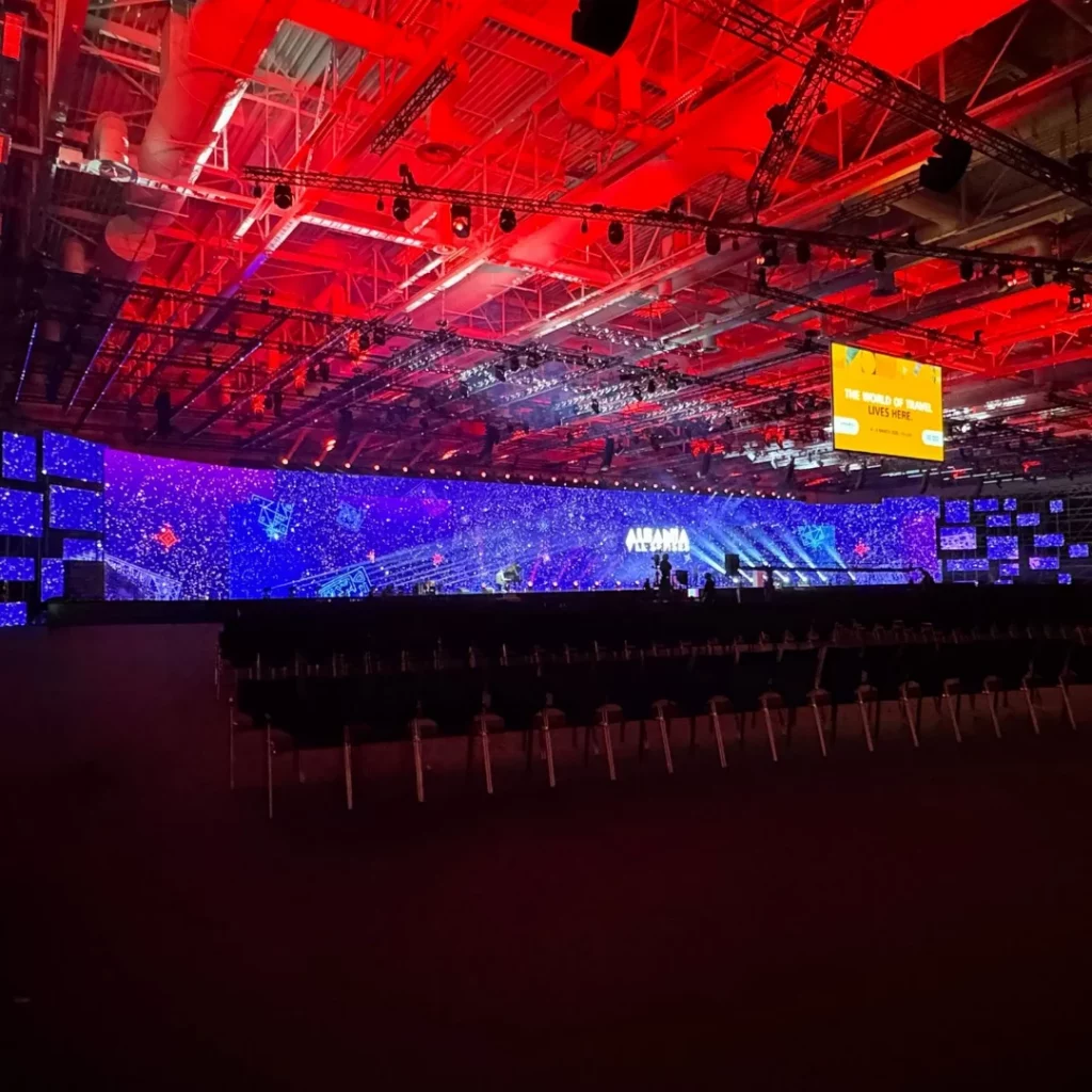 Konferenzhalle mit leeren Stuhlreihen vor Bühne mit blauen und violetten Lichtern, roter Decke und großer Leinwand mit gelber Schrift.