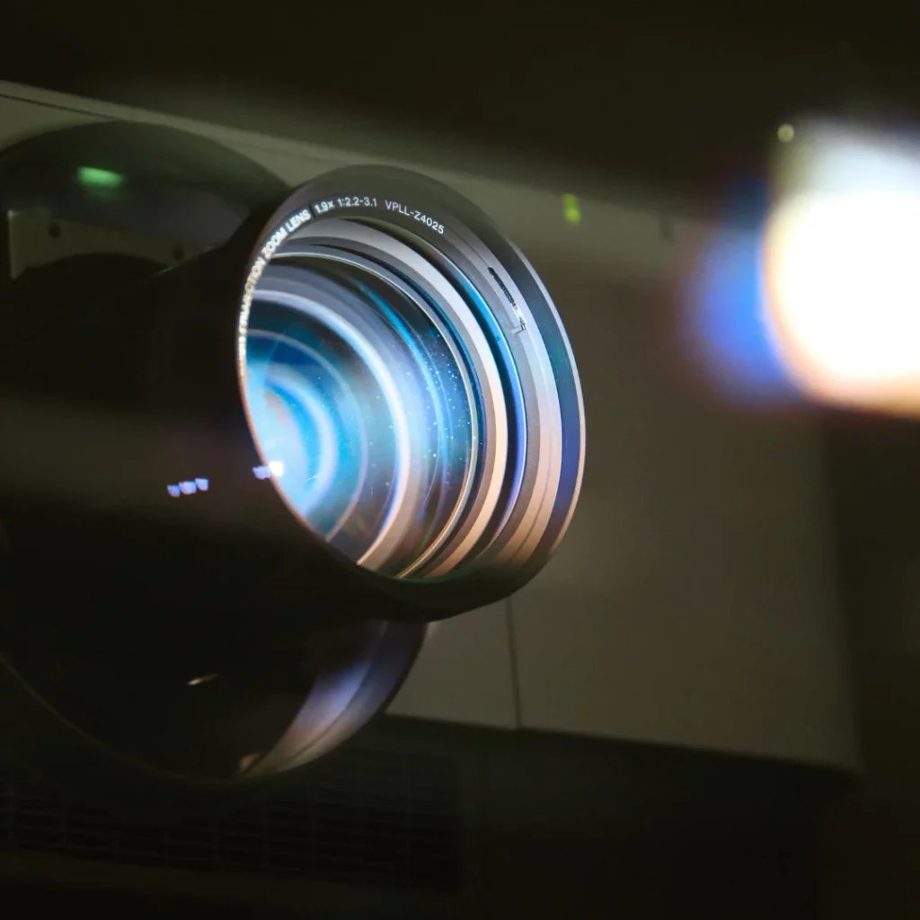 Nahaufnahme eines leuchtenden Beamer-Objektivs mit blauem Lichtreflex, Epson-Projektor in Betrieb, unscharfer Lichtschein rechts.