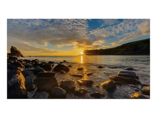 Produktbild eines felsigen Strandes bei Sonnenuntergang: Goldenes Sonnenlicht spiegelt sich auf dem Wasser, darüber ein blauer Himmel mit vereinzelten Wolken, im Hintergrund sanfte Hügel. Ideales Naturmotiv für Wanddekoration oder Postkarten.