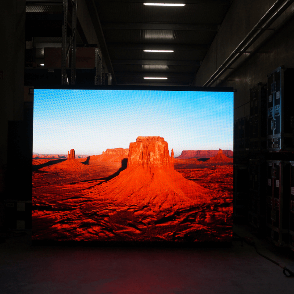 Großes LED-Display in einem schwach beleuchteten Industrie-Raum, auf dem eine leuchtende Wüstenlandschaft mit roten Felsen und blauem Himmel gezeigt wird; das LED-Screen ist für Kauf oder Miete erhältlich.